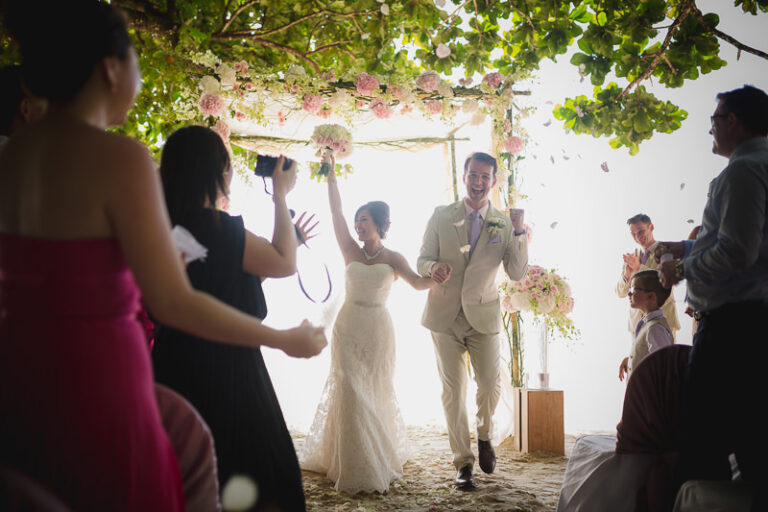 Newlyweds walk back under a flower-hung arch as guests film and confetti falls.
