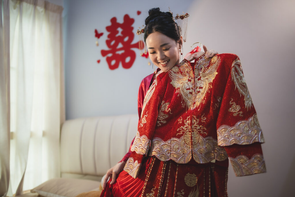 Bride laughing in a red and gold Chinese wedding jacket beside a double-happiness sign.