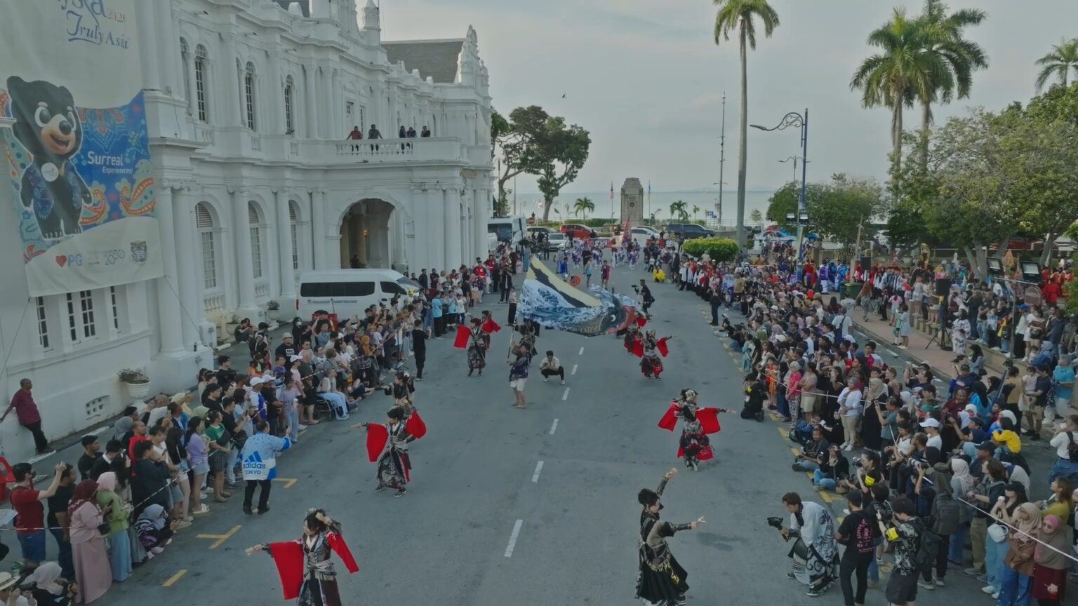 Aerial of the parade filling the road past a white colonial building, crowds either side.