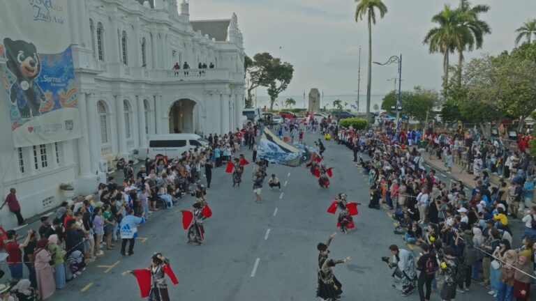 Aerial of the parade filling the road past a white colonial building, crowds either side.